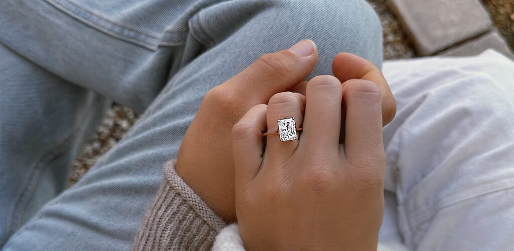 A close-up shot of a couple holding hands, with a diamond engagement ring prominently displayed on one partner's finger, set against a casual outfit background.